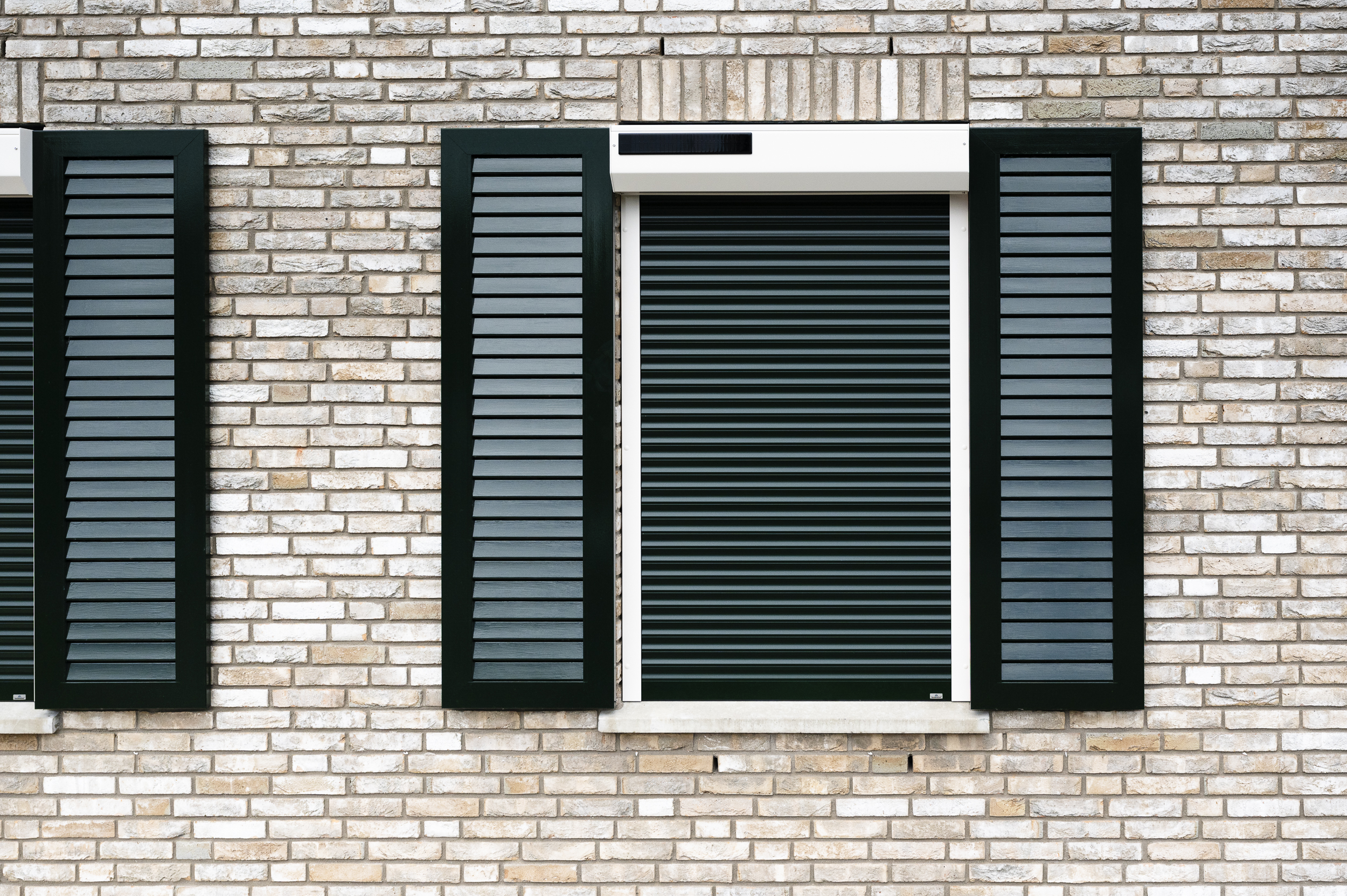 Close-up van een gesloten donkergroen rolluik met ge&iuml;ntegreerd zonnepaneel, omringd door decoratieve groene luiken op een gevel van licht gem&ecirc;leerd metselwerk.