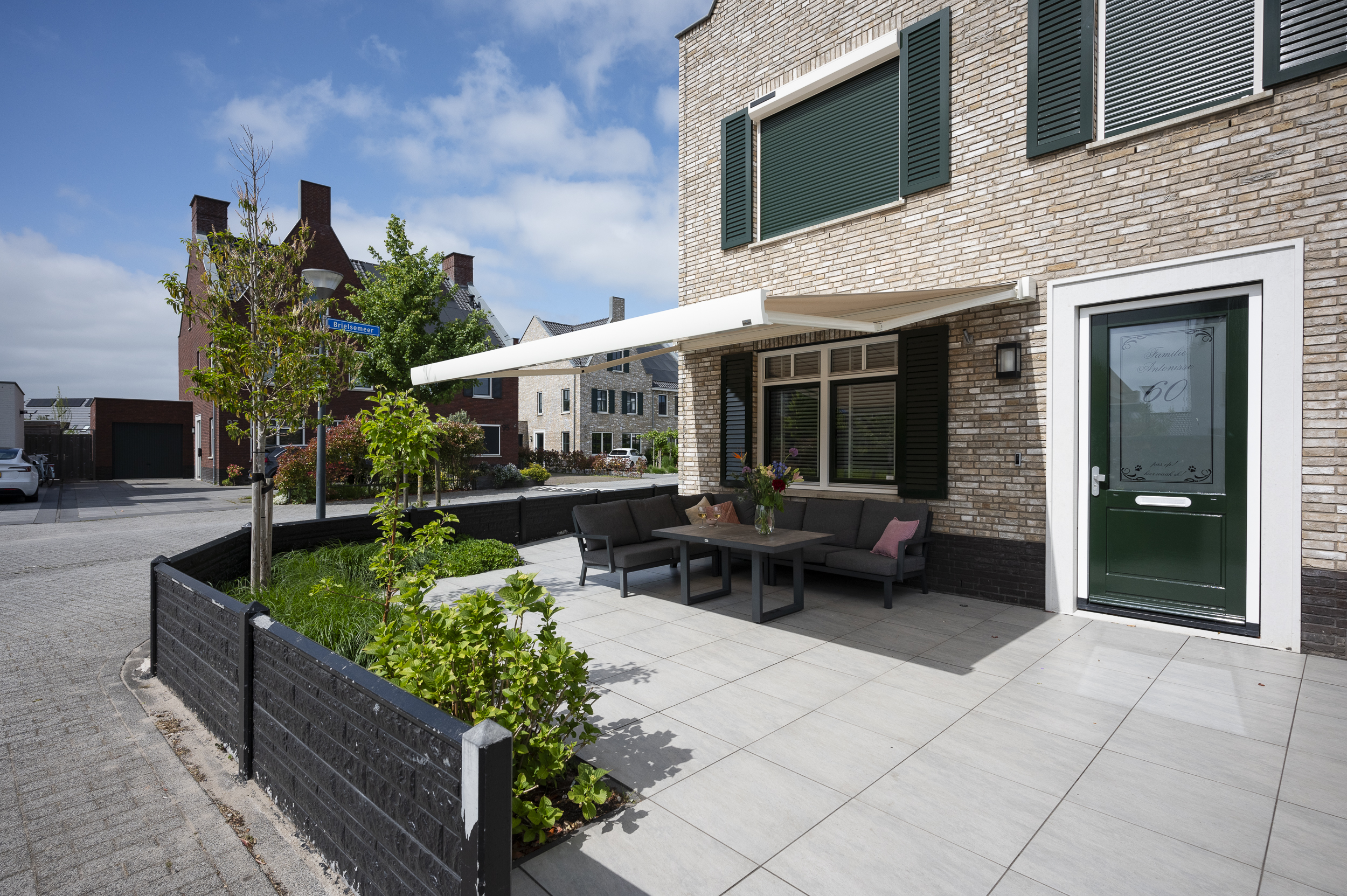 Woning met groene rolluiken en een uitgeschoven wit knikarmscherm boven een modern ingericht terras in een rustige woonwijk in Zoetermeer.